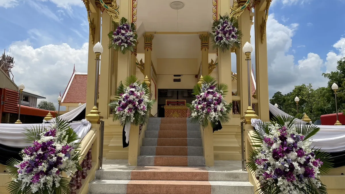 พวงหรีดดอกไม้สดโทนขาวและม่วงอ่อนตั้งเรียงสองข้างทางขึ้นศาลาในงานบำเพ็ญกุศล